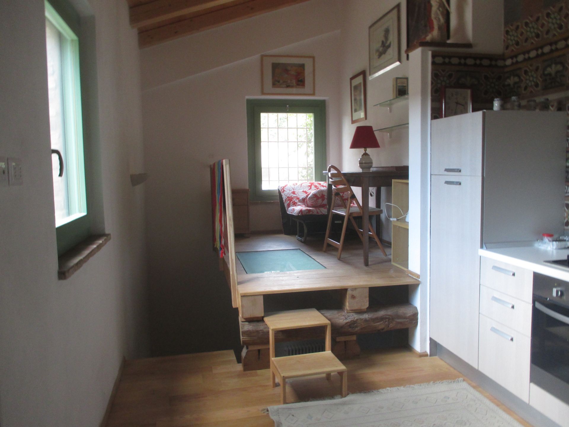 In una stanza moderna, si trova una piccola zona giorno rialzata con scrivania, sedia e divano, accessibile tramite uno sgabello di legno.