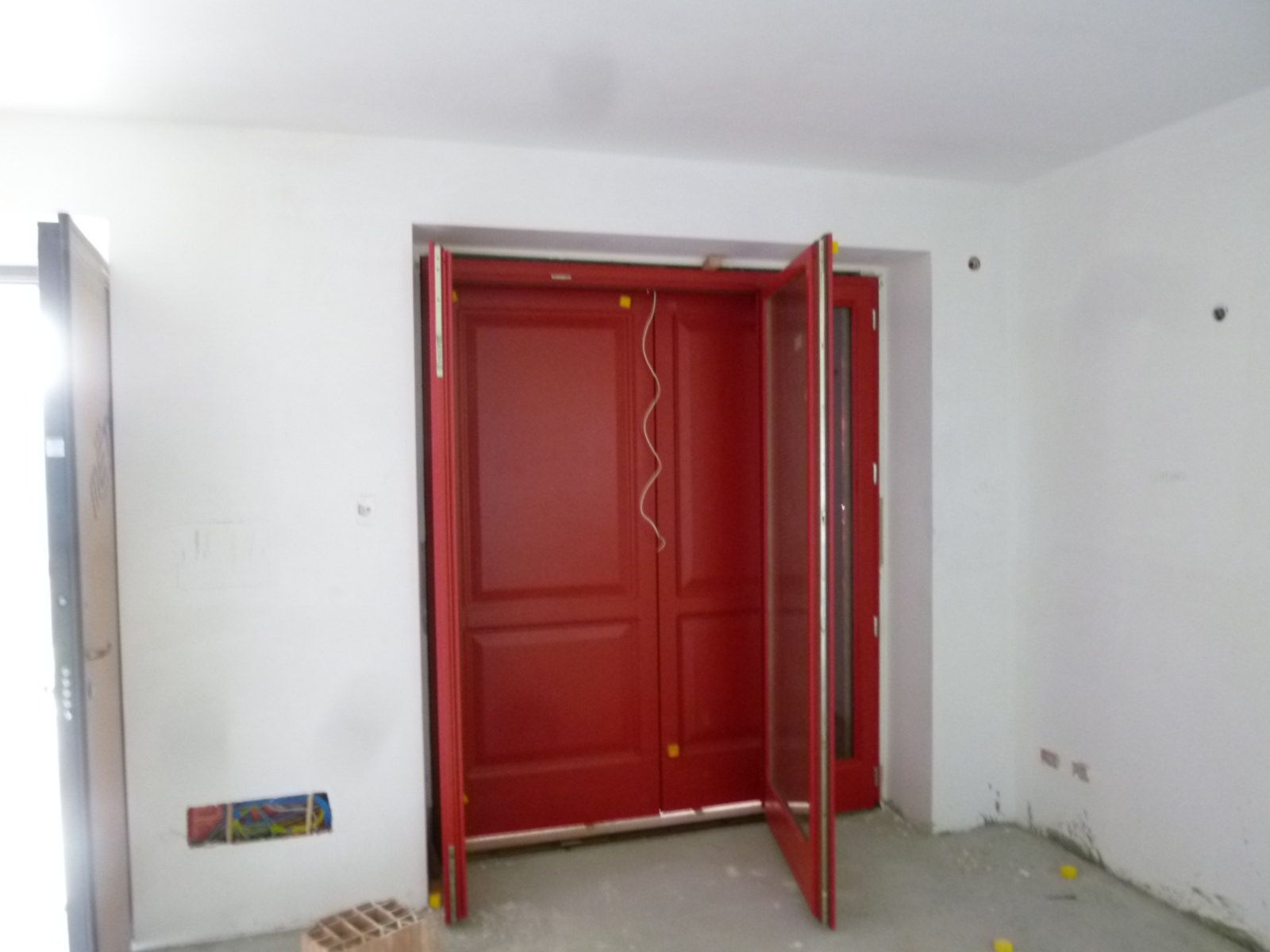 Una stanza in fase di ristrutturazione con pareti bianche e una grande porta a due ante rossa al centro dell'ingresso.
