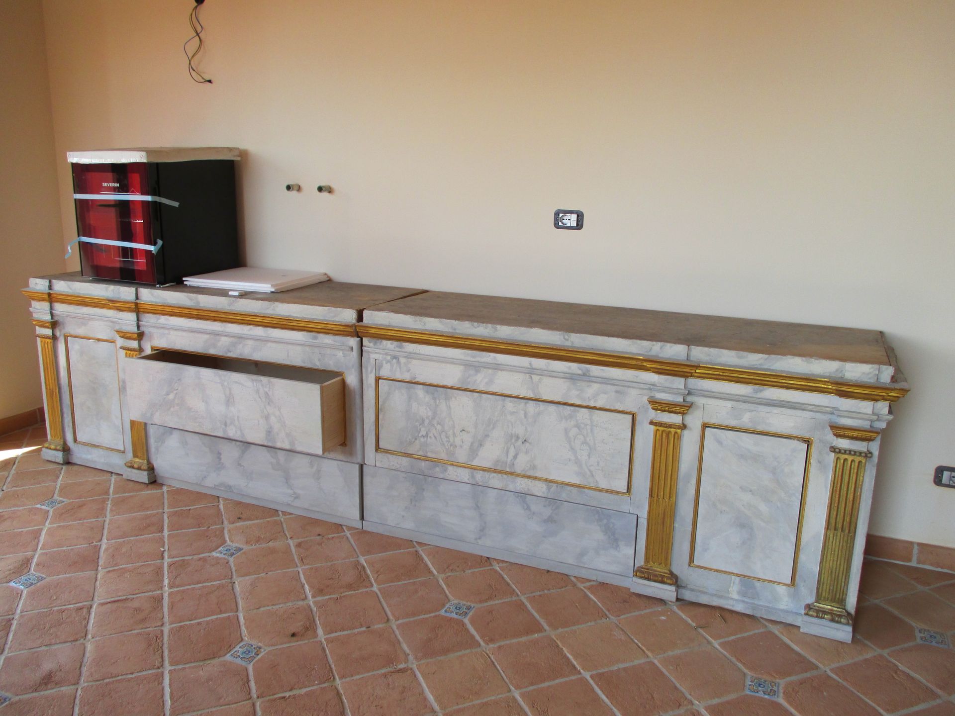 Una lunga credenza con piano in marmo, finiture e pilastri dipinti d'oro è addossata a una parete spoglia, su un pavimento piastrellato.