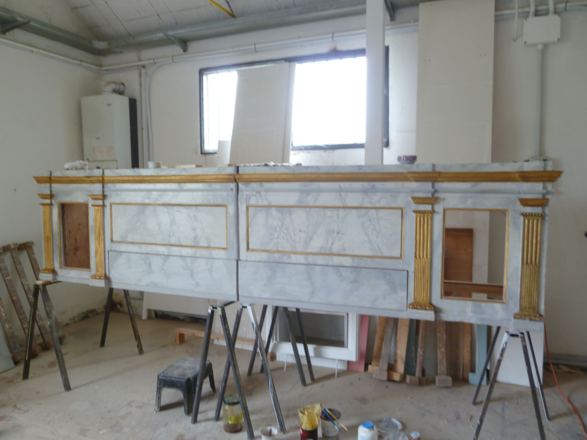 In un'officina, due sezioni di un altare in legno con finiture dorate e rivestimento in finto marmo poggiano su cavalletti.