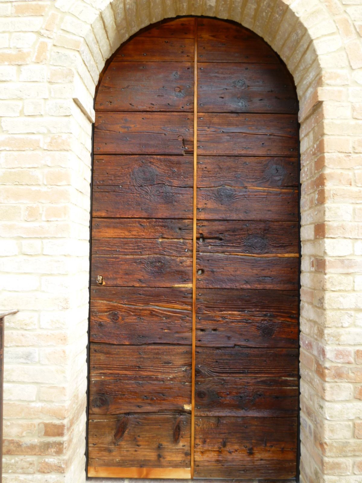 Una porta a doppio battente in legno rustico, tinta di scuro, inserita in un ingresso ad arco in mattoni chiari.