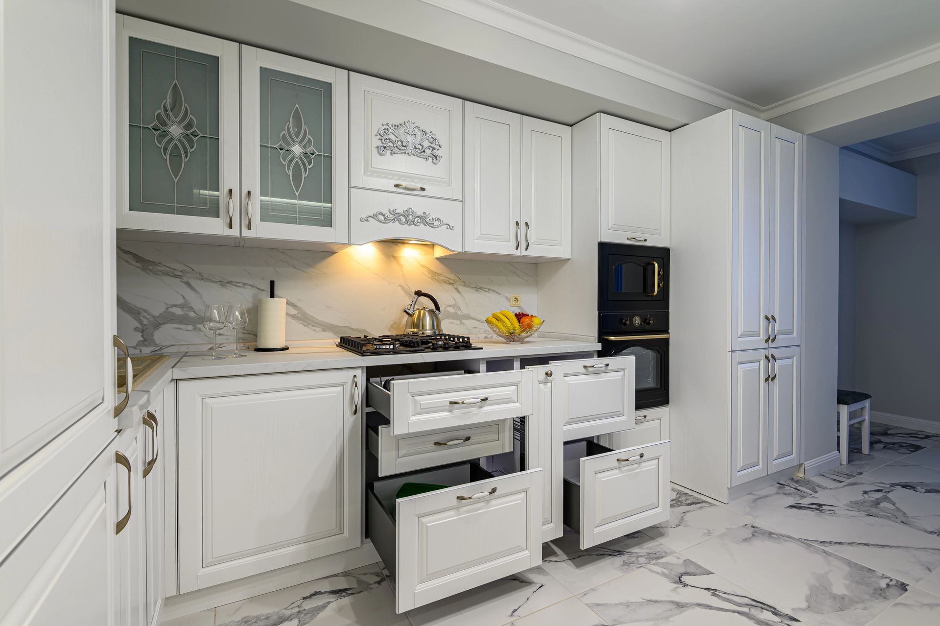 Una cucina luminosa con mobili bianchi, paraschizzi e pavimento effetto marmo, dotata di un mobile contenitore con cassetti a vista.