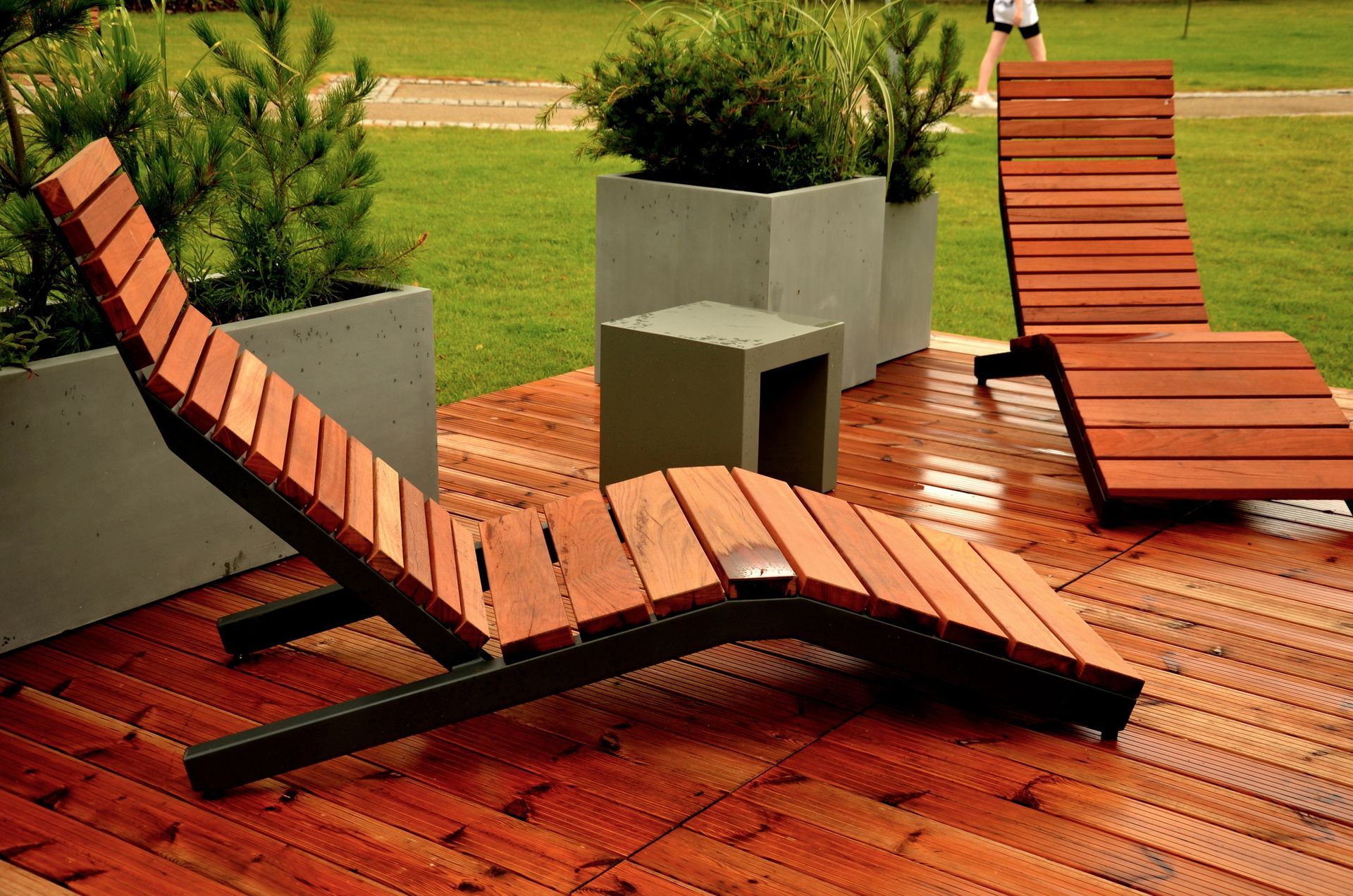 Due moderne sedie a sdraio con struttura a doghe di legno su una terrazza in legno con fioriere in cemento grigio, sullo sfondo di un prato verde.