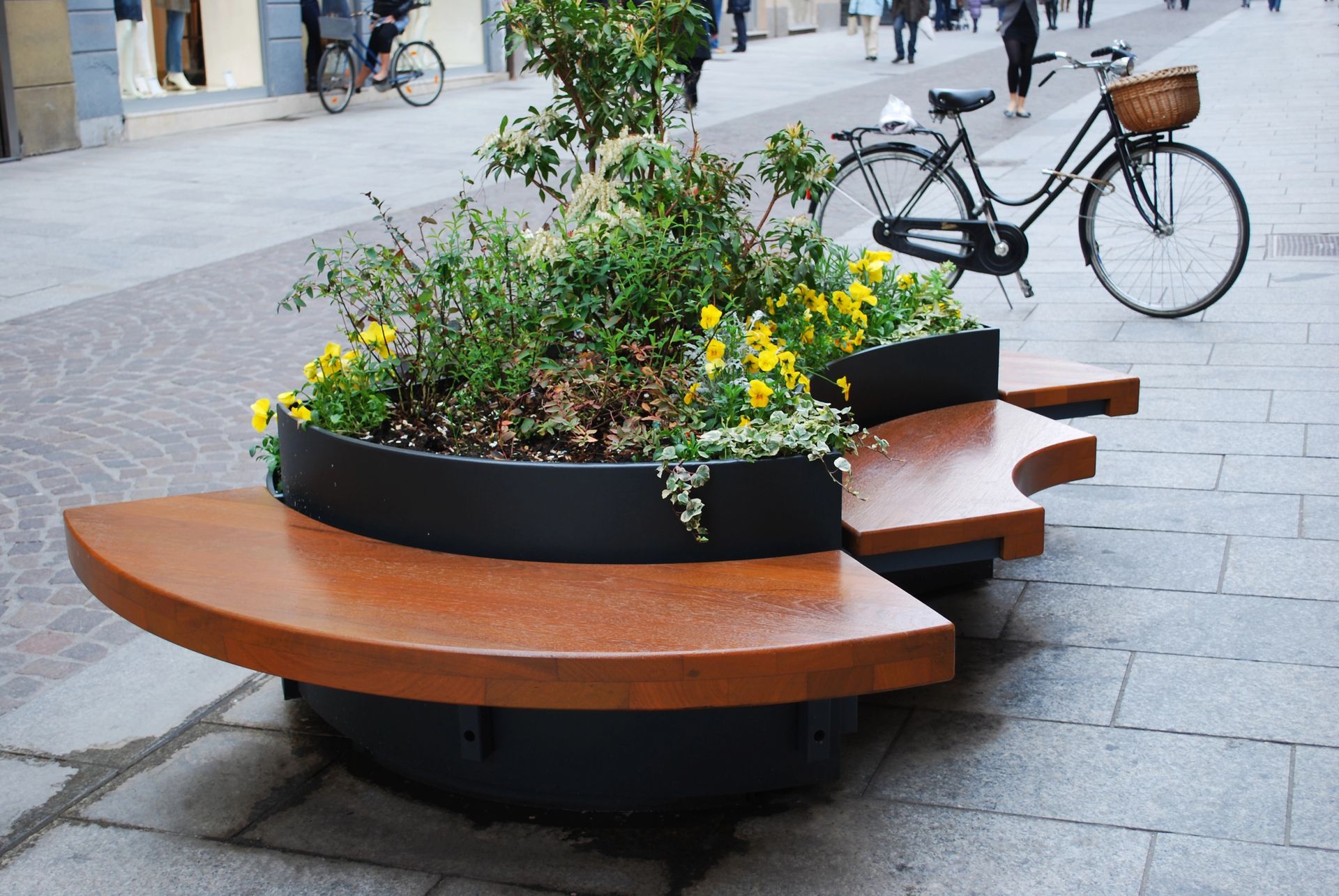Una fioriera circolare con panchine in legno su una via pedonale, con una bicicletta parcheggiata sullo sfondo.