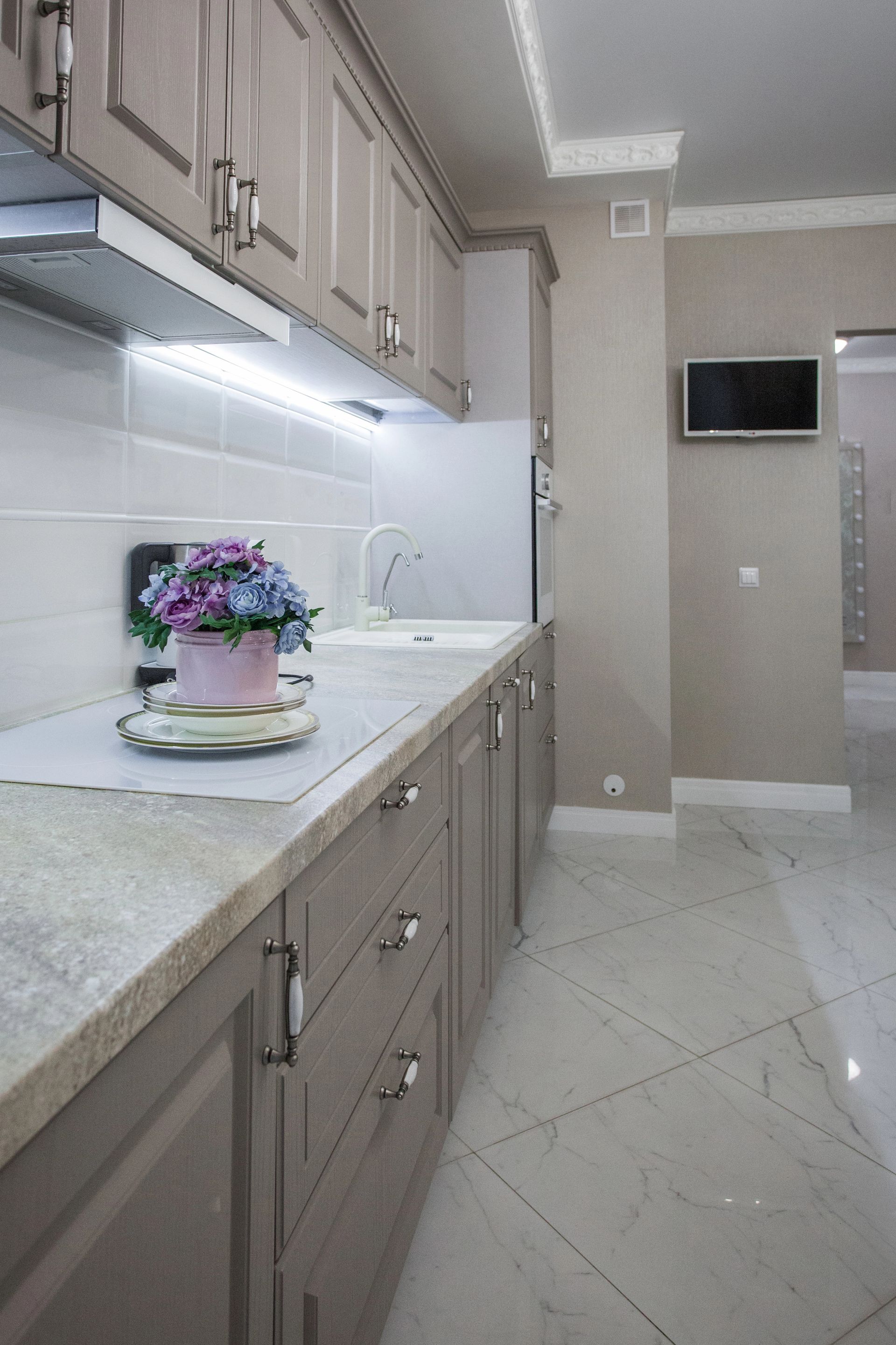 Cucina moderna con mobili color tortora, ripiani in pietra chiara, paraschizzi in piastrelle bianche e decorazioni floreali.