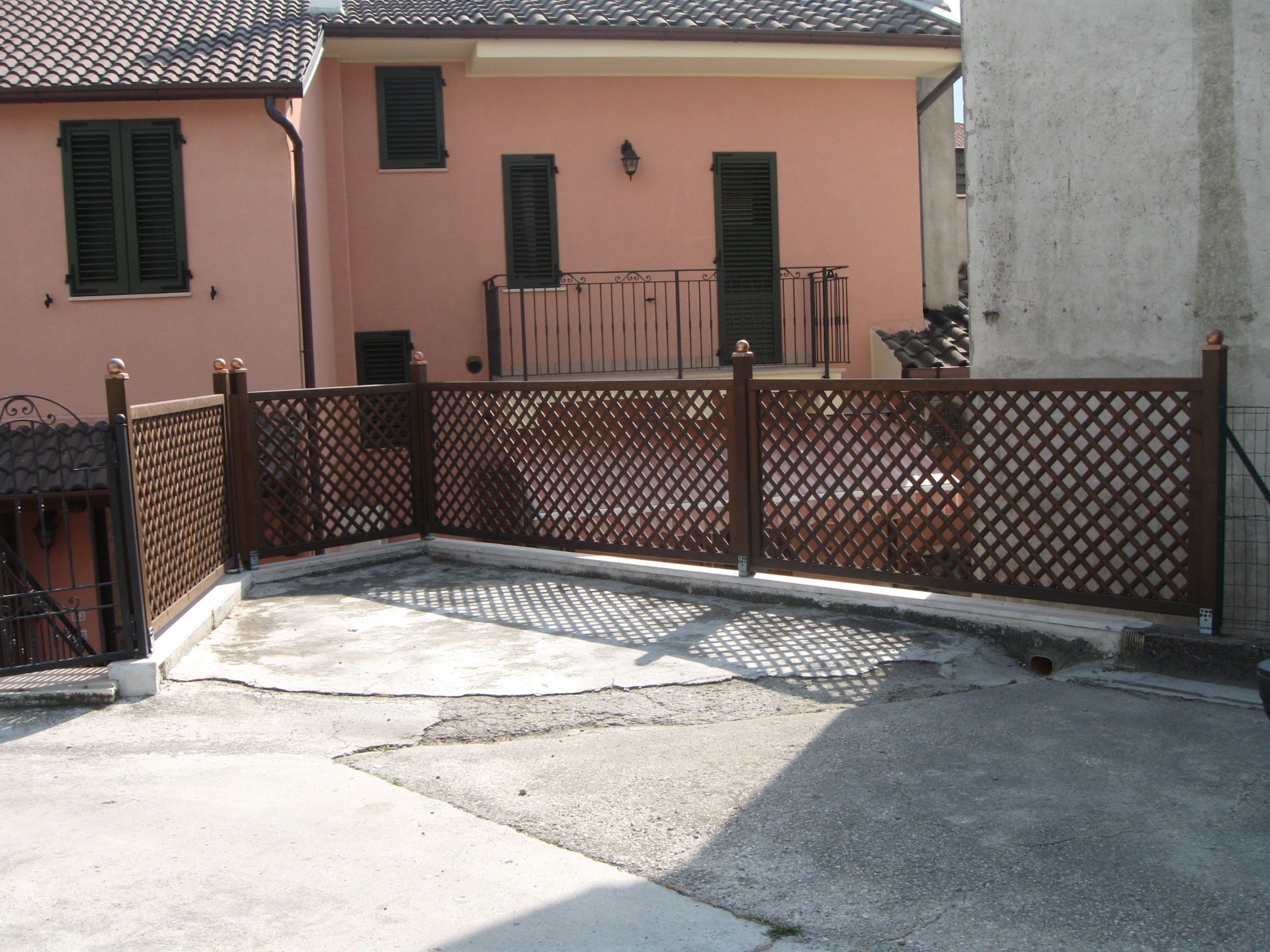 Una recinzione a traliccio in legno marrone si erge su un patio in cemento di fronte a una casa a due piani di colore rosa chiaro.