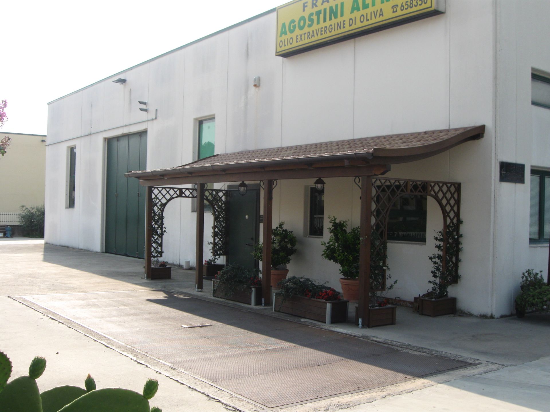 Un edificio commerciale con esterni bianchi, un grande portone a bovindo verde e un portico in legno marrone ricoperto di vegetazione.