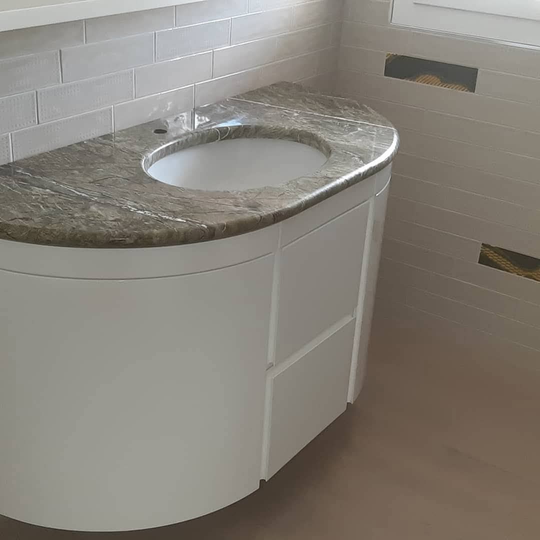 Un mobile da bagno curvo di colore bianco con piano e lavabo in granito, installato a ridosso di una parete piastrellata.