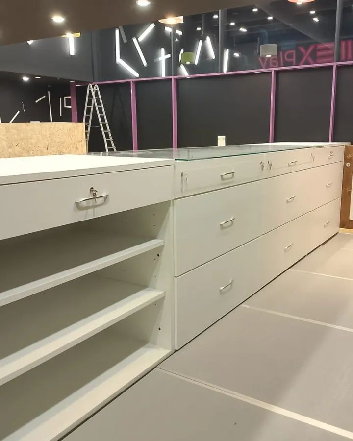 Un lungo bancone bianco con cassetti e ripiani a giorno in un negozio in costruzione, con una scala sul retro.