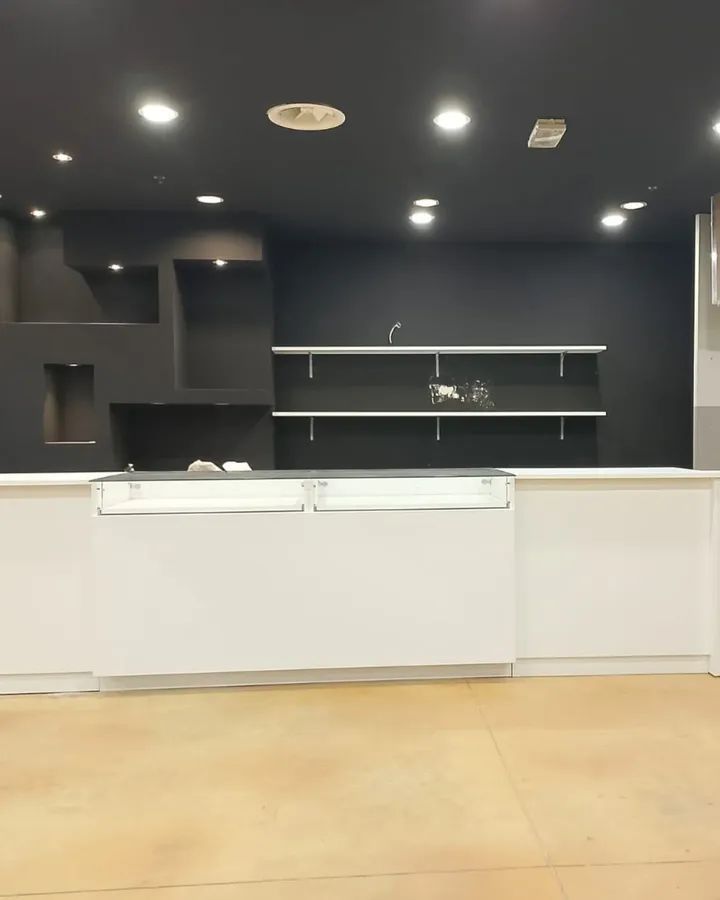 Interno di un negozio moderno caratterizzato da un bancone espositivo bianco di fronte a una parete scura con scaffali e illuminazione integrati.