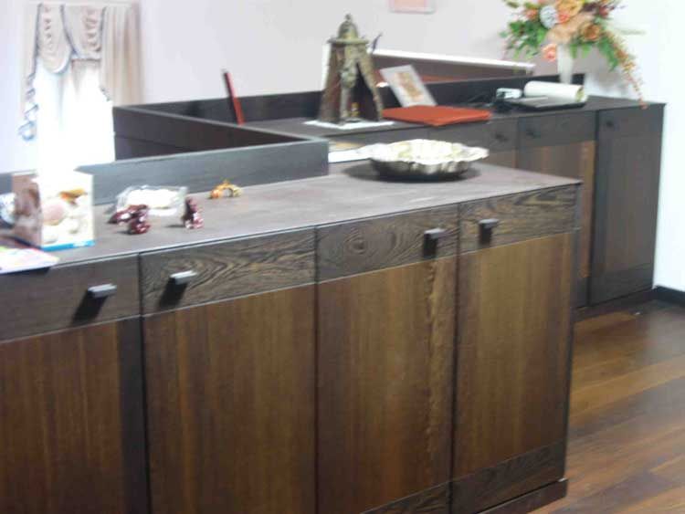 Vista di un bancone reception in legno scuro con cassetti, oggetti decorativi e una ciotola sul ripiano in un ufficio professionale.