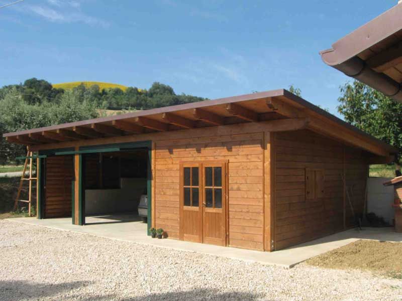 Un garage e un capanno per attrezzi in legno con tetto spiovente e doppie porte, situati su un terreno ghiaioso sotto un cielo azzurro e limpido.