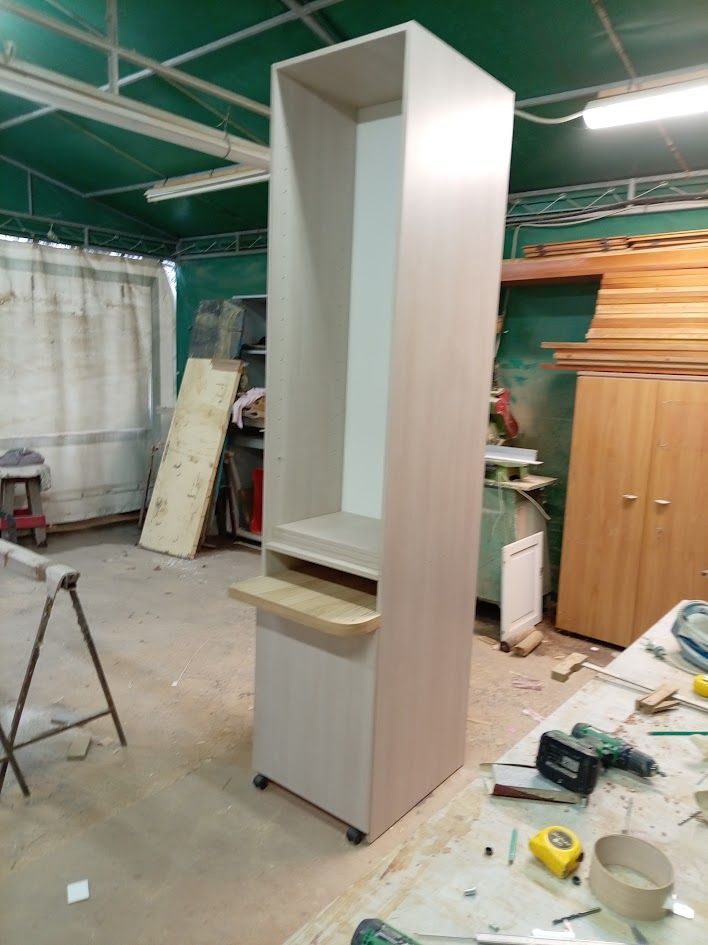 Un armadio alto, di colore legno chiaro, su ruote, con un ripiano superiore aperto e un piccolo piano pieghevole, in un laboratorio di falegnameria.