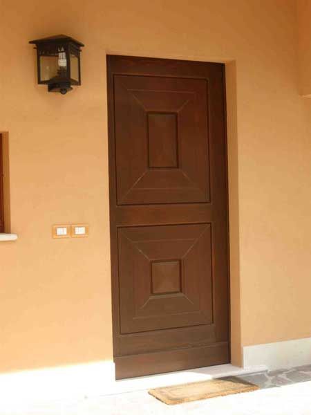 Una porta in legno a due pannelli, di colore marrone scuro e con un motivo geometrico, incastonata in una parete beige chiaro accanto a una lampada da parete per esterni.