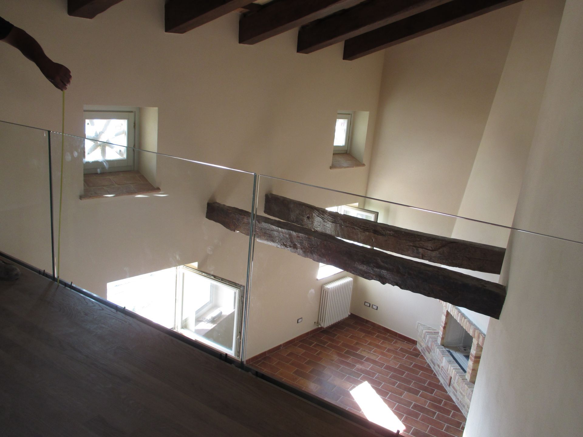 Una vista dall'alto da un soppalco con ringhiera in vetro che si affaccia su una zona giorno rustica con travi a vista sul soffitto.