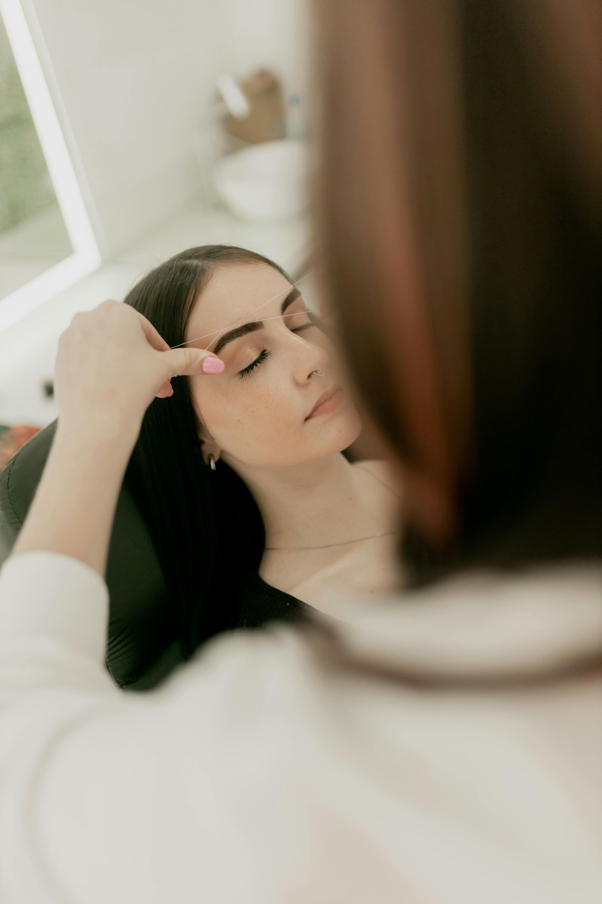 Woman receiving eyebrow treatment in a well-lit salon; technician touching brow, patient with eyes closed.