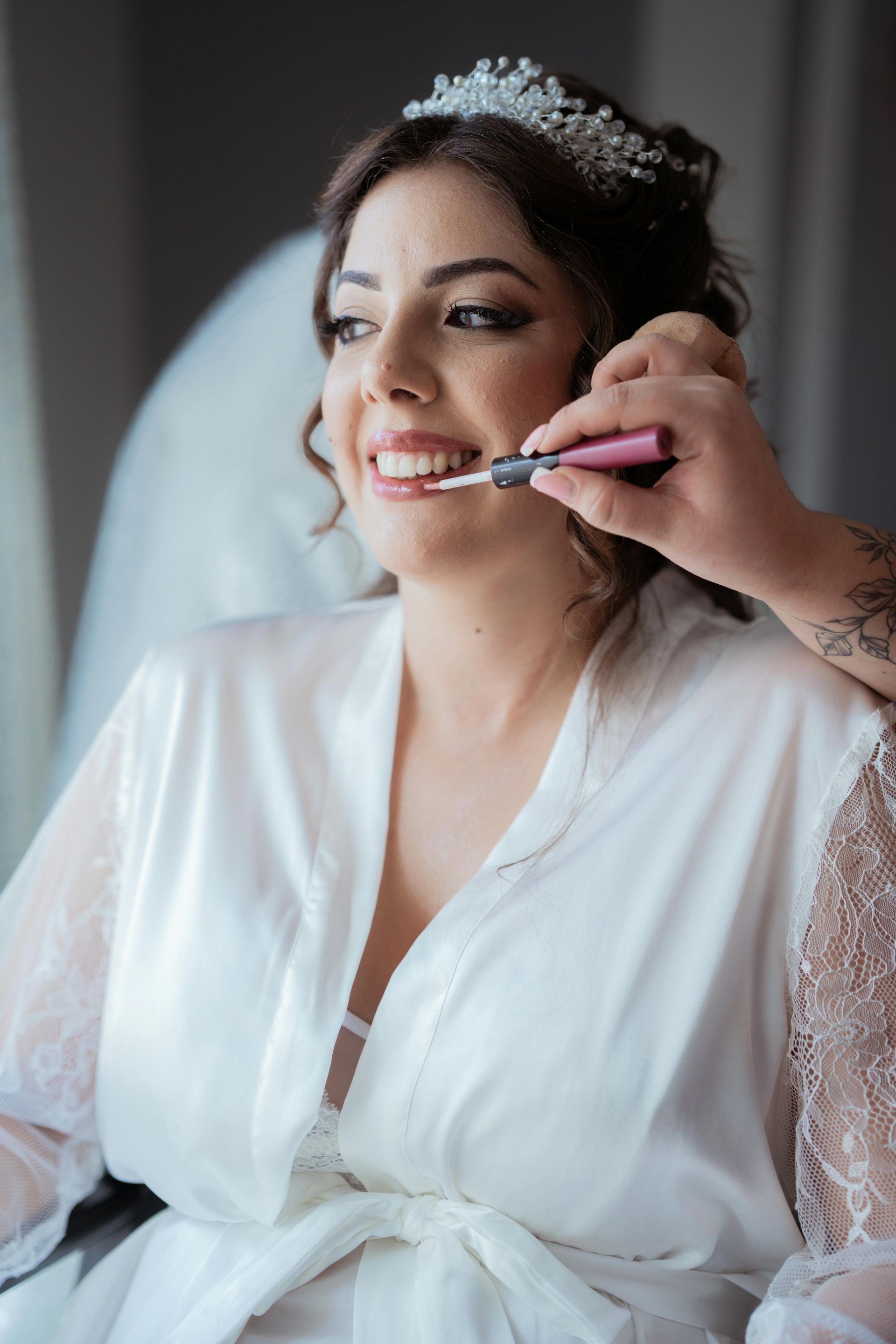 Bride getting lipstick applied, wearing a white robe and tiara, by a window.