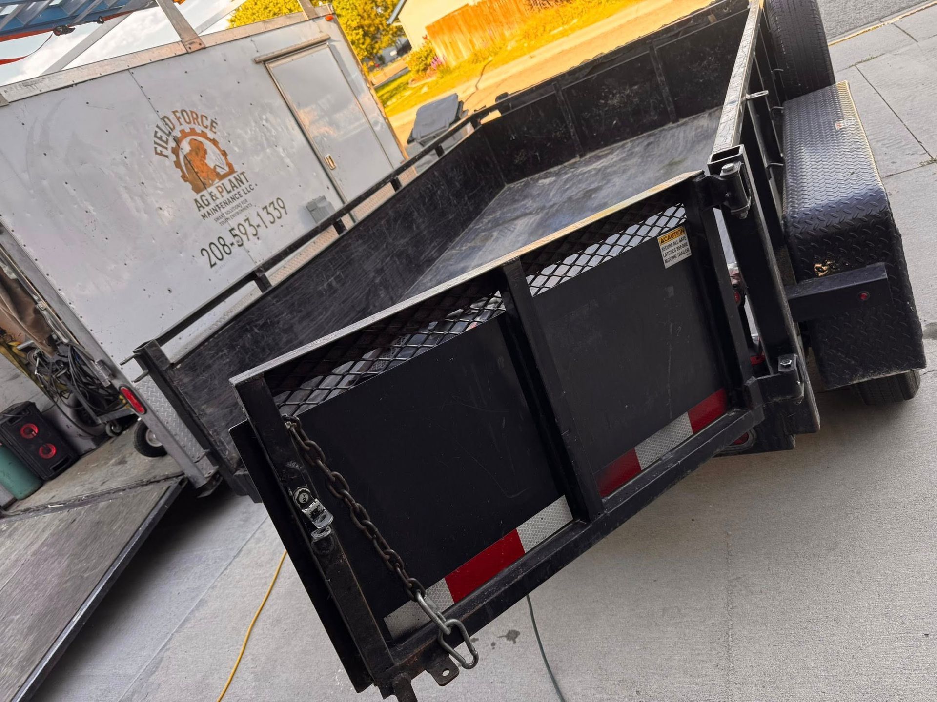 Black utility trailer with side rails parked on pavement, next to a white truck.