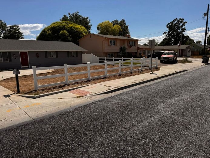 Residential street view: A white fence surrounds a small yard in front of a gray and tan building on a sunny day.