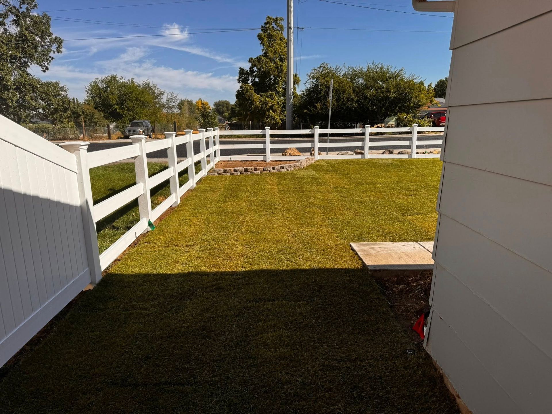 White fence encloses a green lawn. Sunlight casts shadows on the grass. A house corner is on the right.