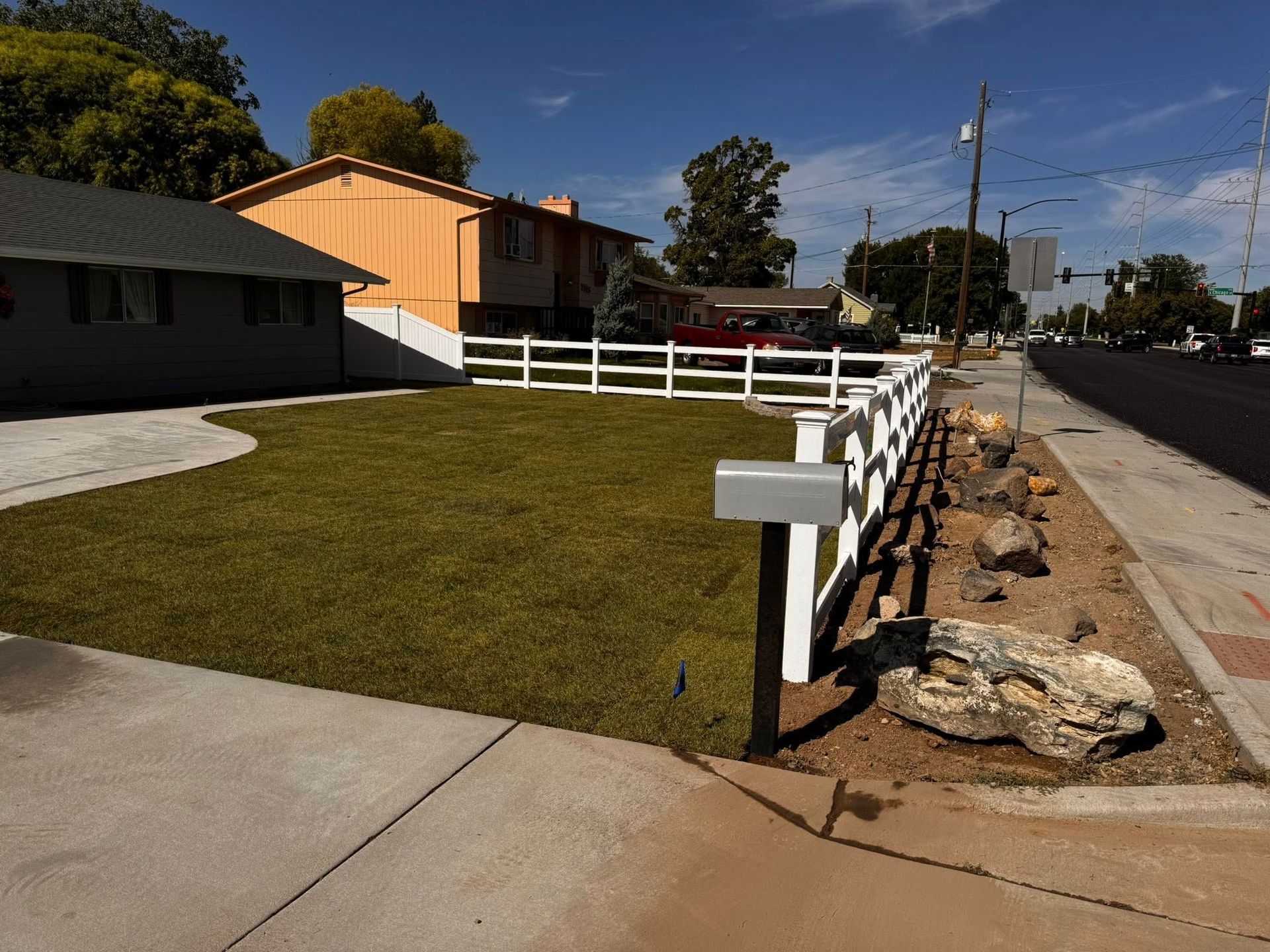 House with white fence, green lawn, and mailbox next to a street.