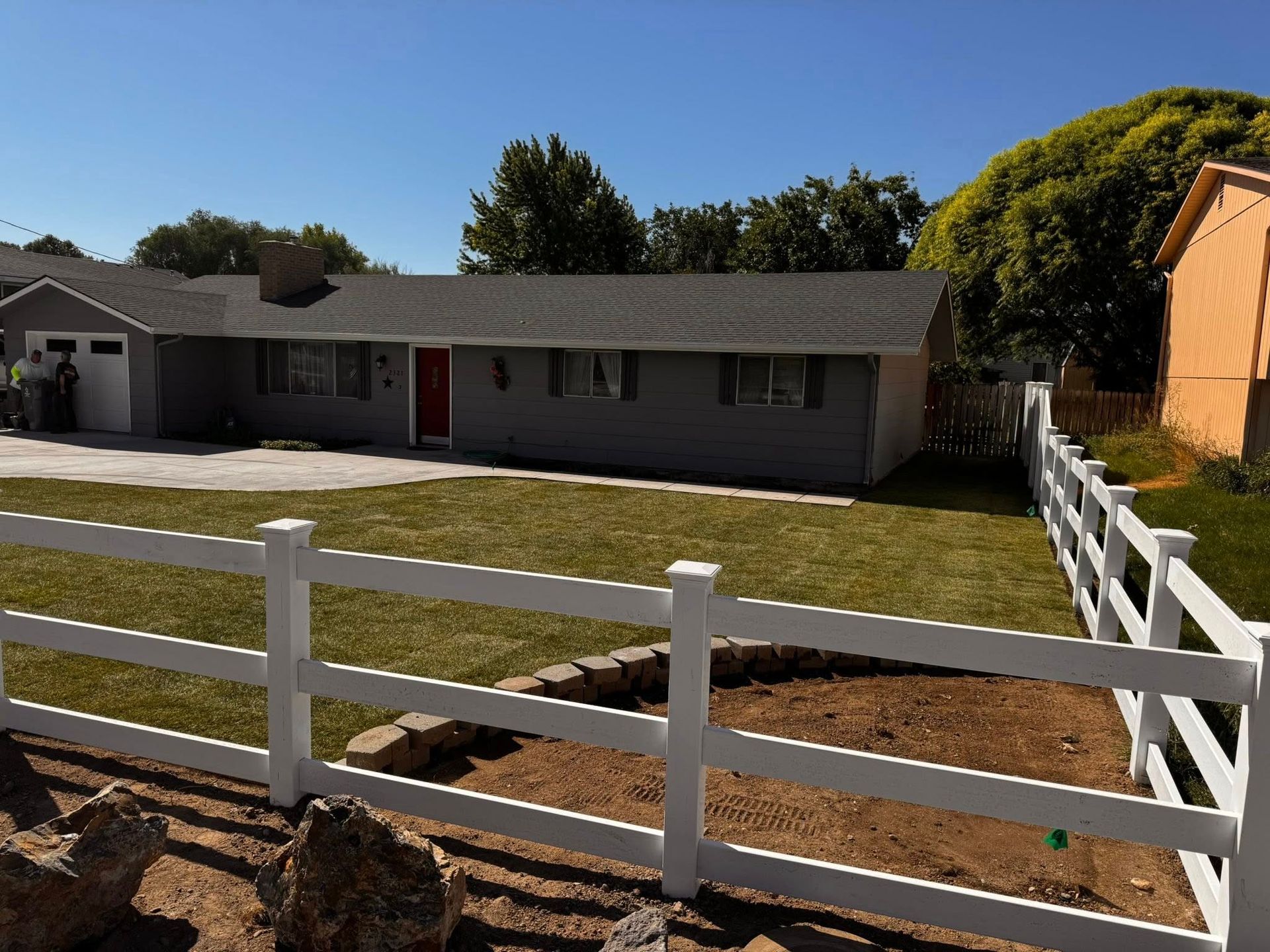 Gray house with a white fence and a red door. Green lawn under a blue sky.