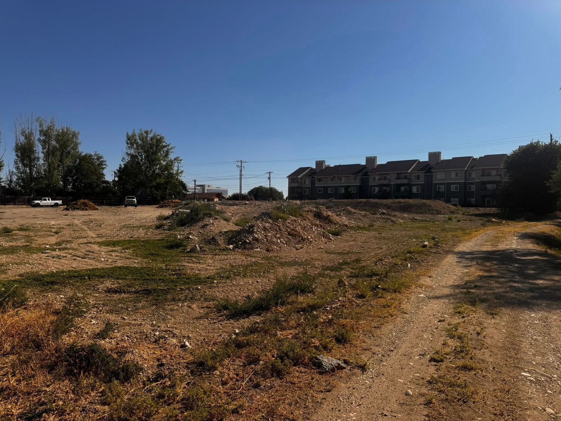 Dirt field with low vegetation, adjacent to a dirt path, with buildings and trees under a blue sky.