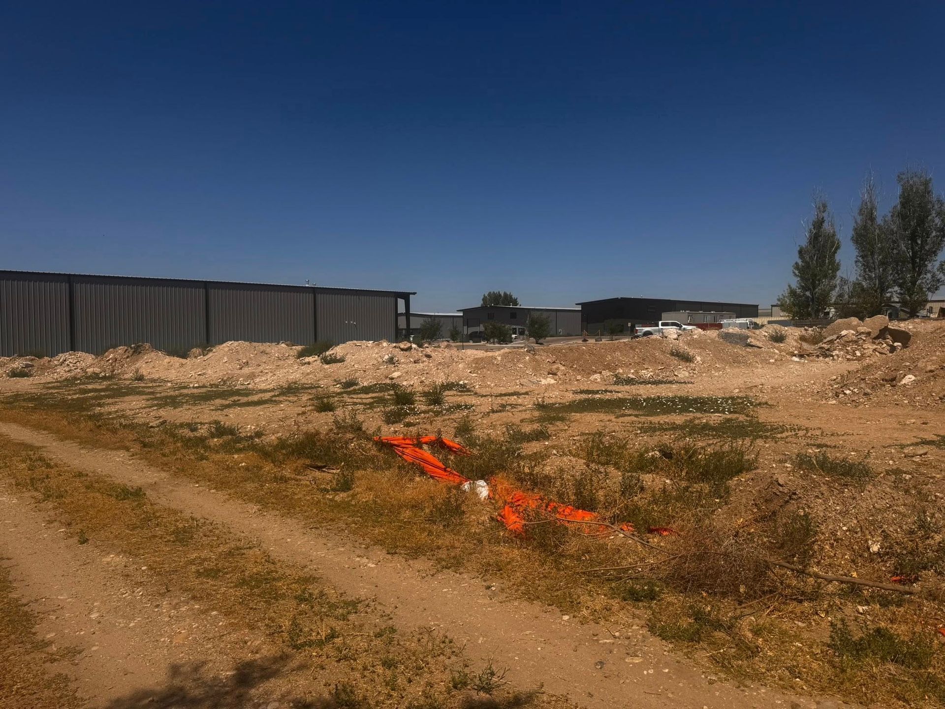 Dirt road leads to construction site under blue sky. Orange marking on ground, structures in background.