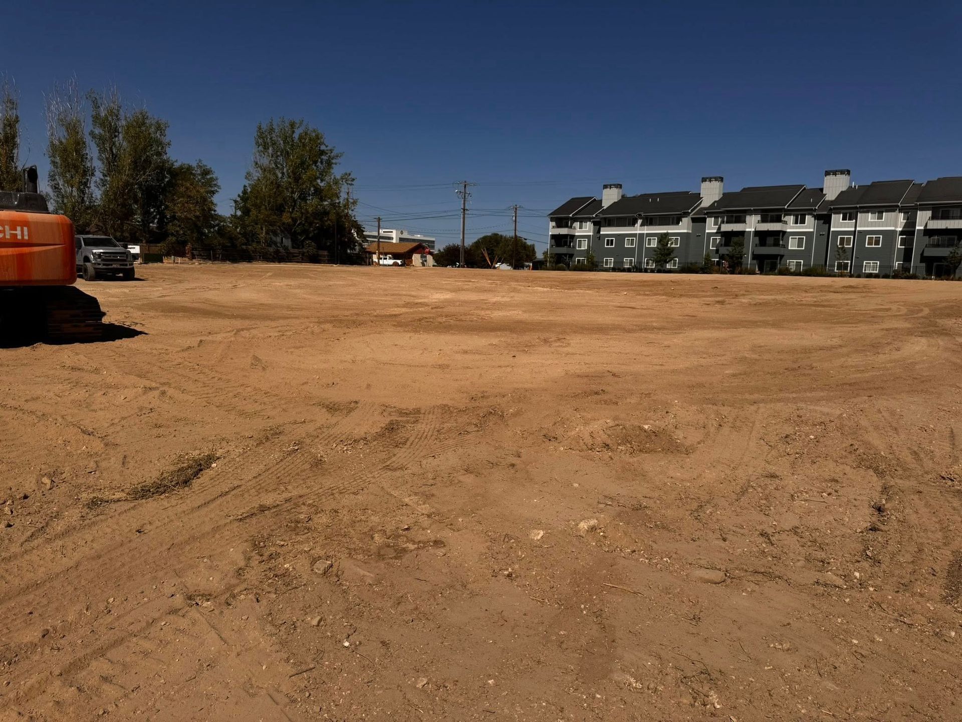 Cleared dirt lot with construction equipment and apartment buildings in the background under a blue sky.