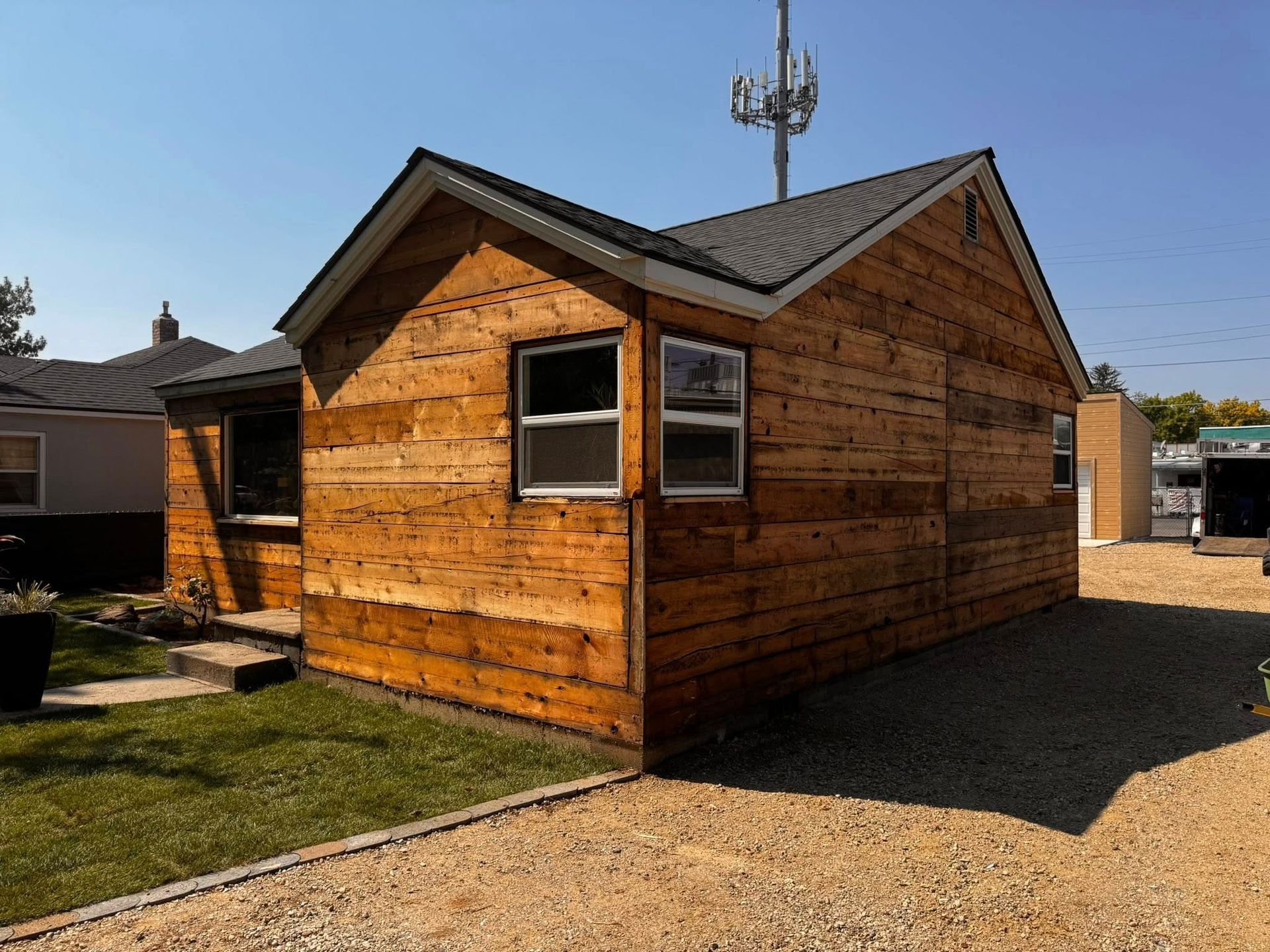 Wooden shed with windows, dark roof, and gravel driveway on a sunny day.