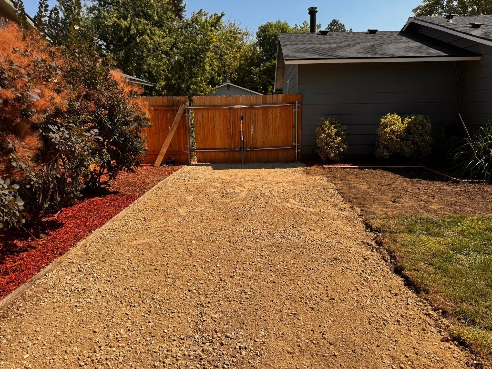 Gravel driveway leads to a wooden gate and house. Red mulch and grass border the driveway. Sunny, outdoor setting.