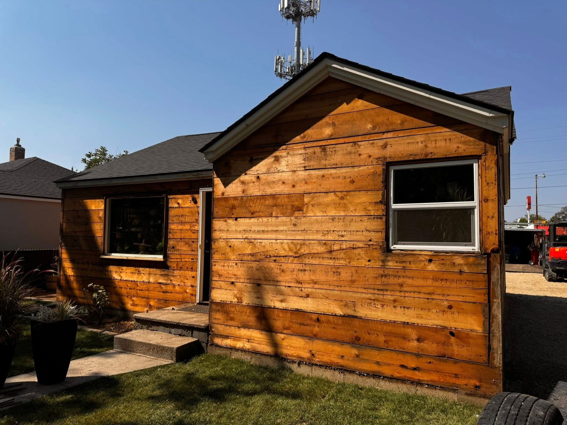 Small wooden building with two windows, dark roof, and concrete steps on a sunny day.