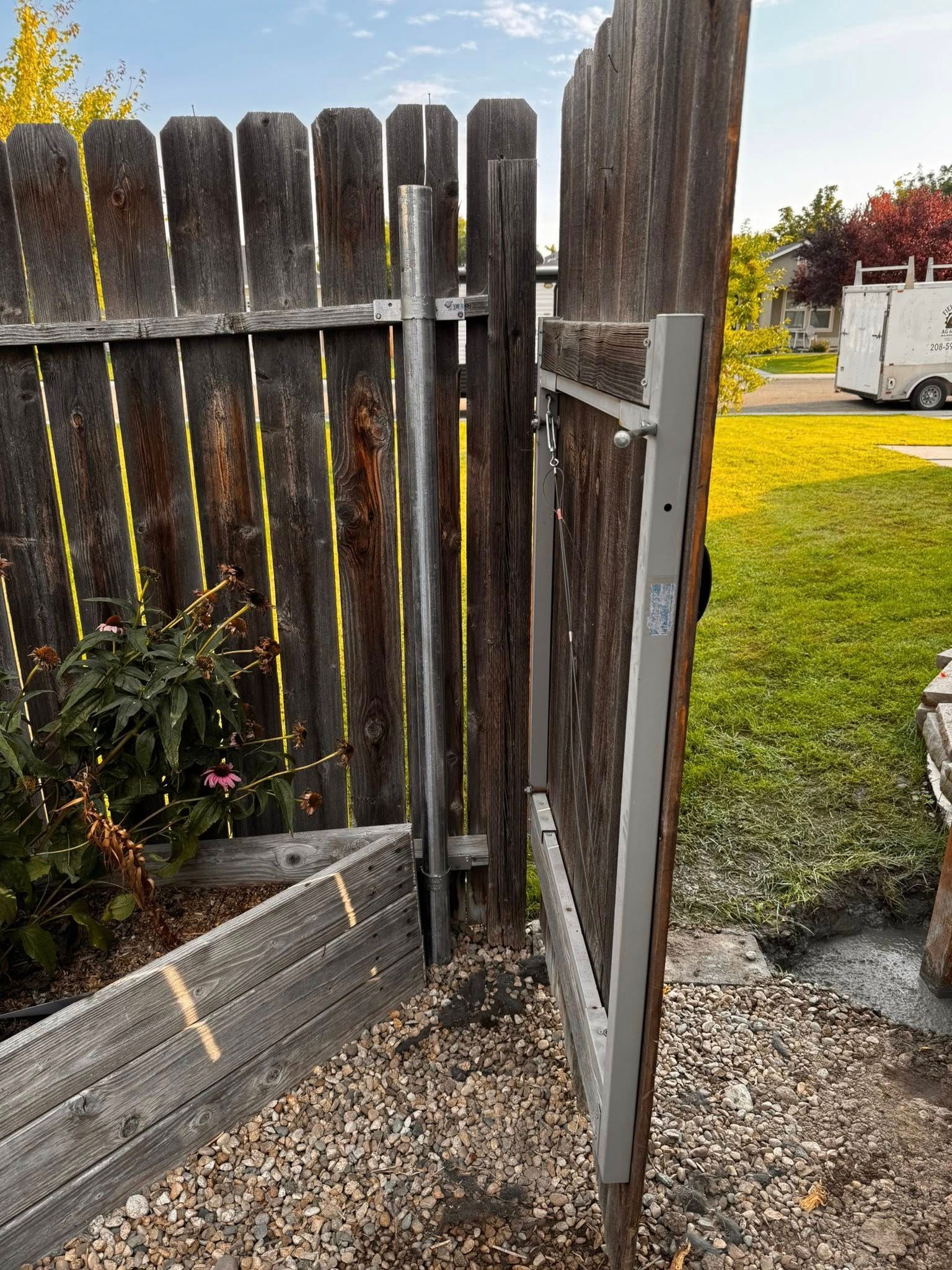 Wooden gate ajar in a weathered fence, gravel ground, and lush greenery in the background.