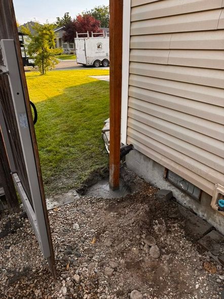 Brown post cemented by beige siding. Dirt, gravel, and a fence are in the foreground.