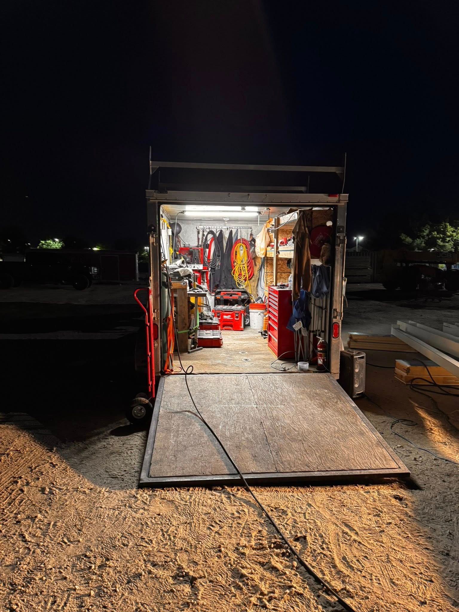 Trailer interior at night, lit up, filled with tools and equipment. Ramp down, red tool chests, workbench visible.