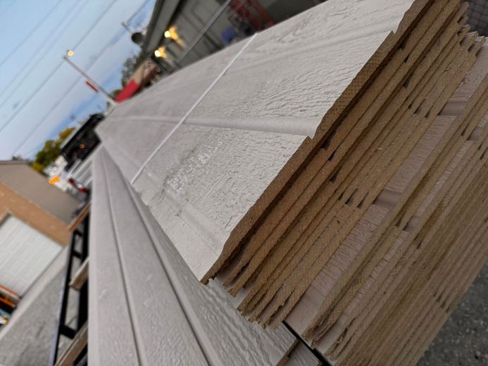 Pile of beige wood siding, textured, stacked outdoors.