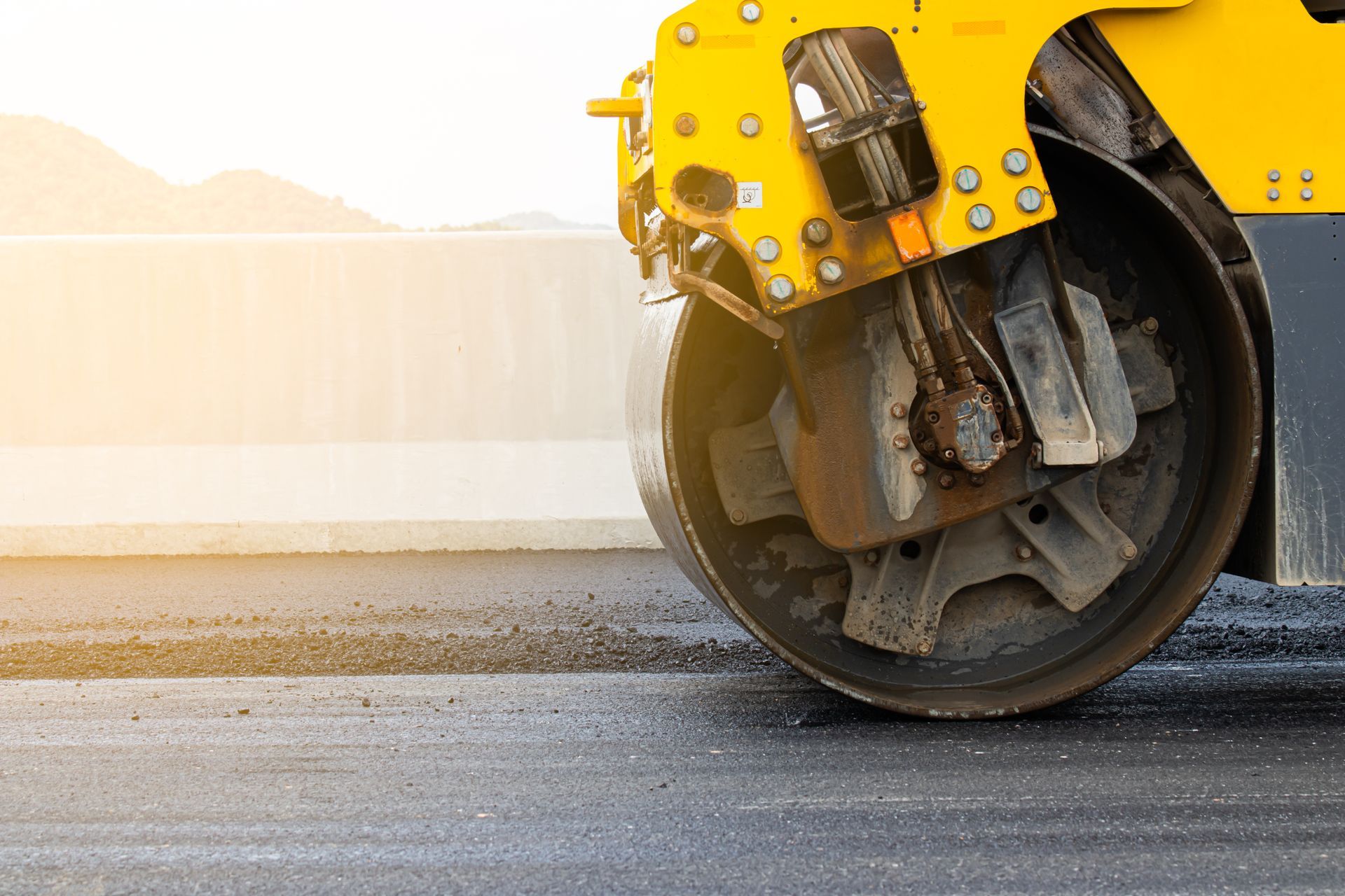 Yellow road roller smoothing wet pavement, with steam rising from the freshly paved surface. Yellow road roller compactor on a paved surface at sunrise, with the front drum in view