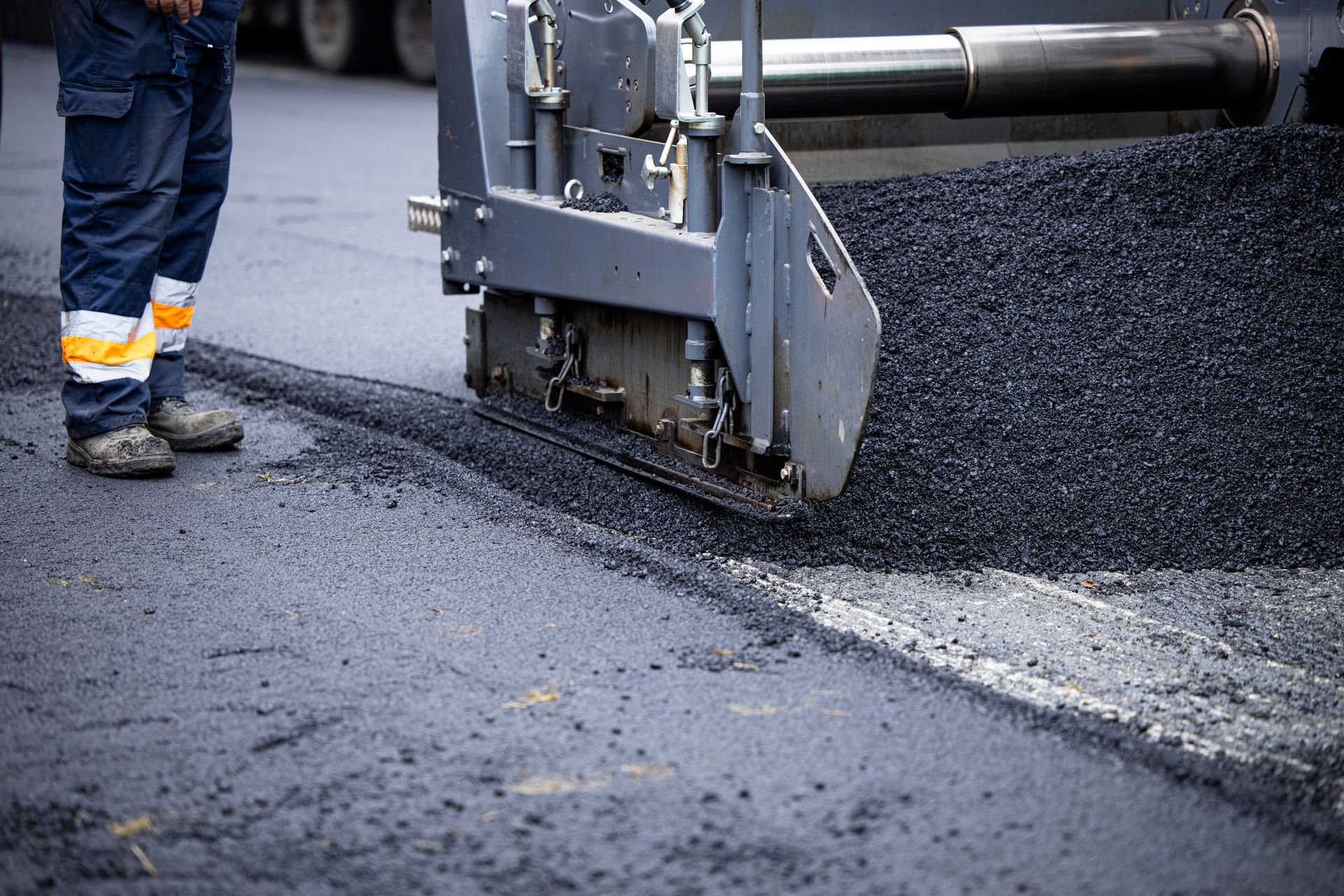 Road paving machine laying fresh black asphalt on a street beside a worker's boots Road paving machine laying fresh black asphalt on a street beside a worker's boots