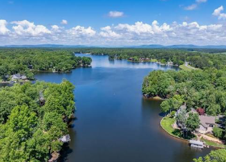 Aerial view of a calm blue lake surrounded by dense green trees and a few lakeside homes under a bright sky