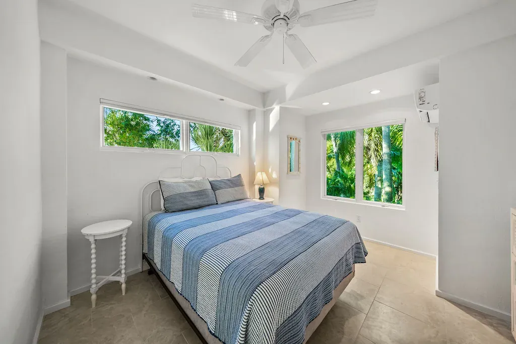 White bedroom with bed, blue bedding, windows, and a small table.