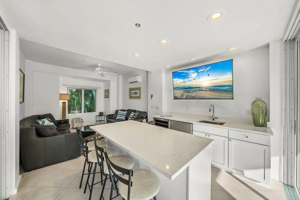 Bright, modern kitchen with island seating. A TV is mounted above the sink and cabinets, with a view of the beach.