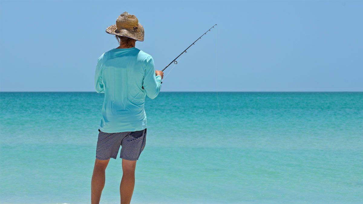 Man in hat and blue shirt fishing in turquoise ocean, sunny day.