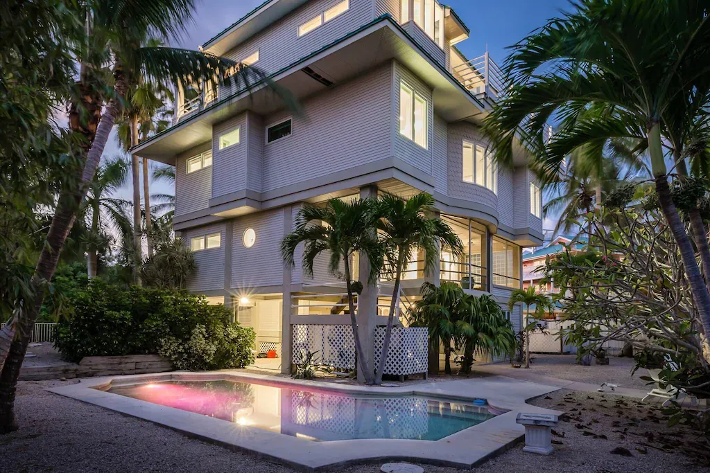 Three-story white house with a pool, palm trees, and a lit-up pool at dusk.