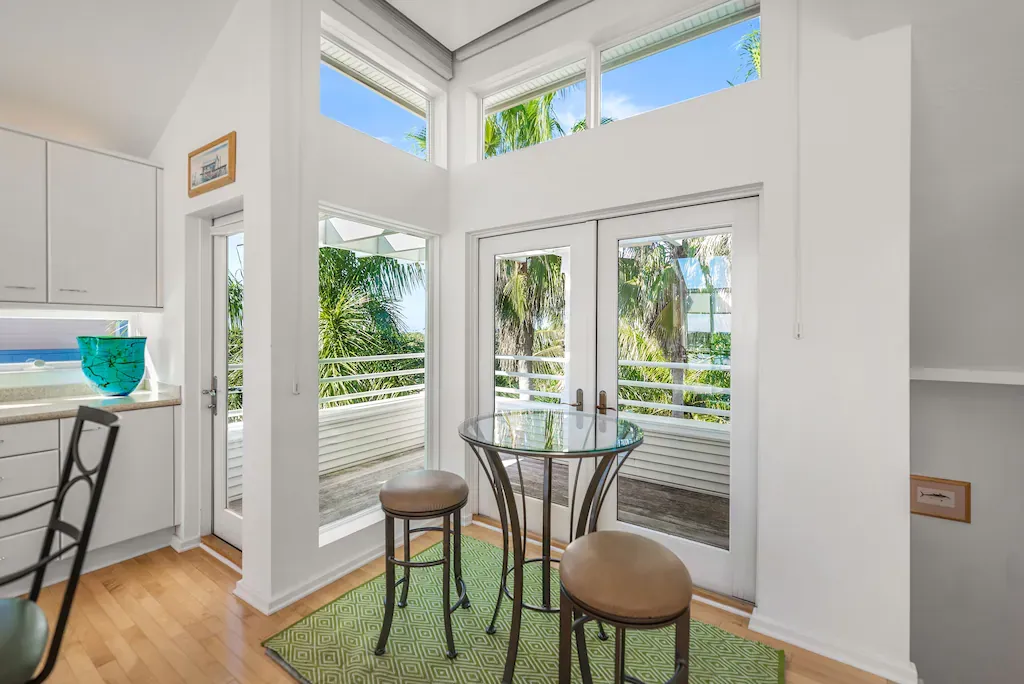 Bright kitchen with high windows, a small table, and stools, overlooking a balcony with trees.