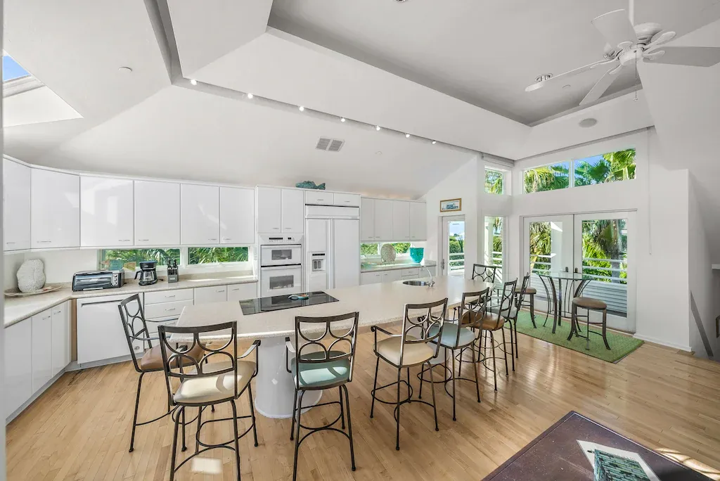 Spacious all-white kitchen with a large island, high ceilings, and wooden floor, overlooking a deck with green plants.