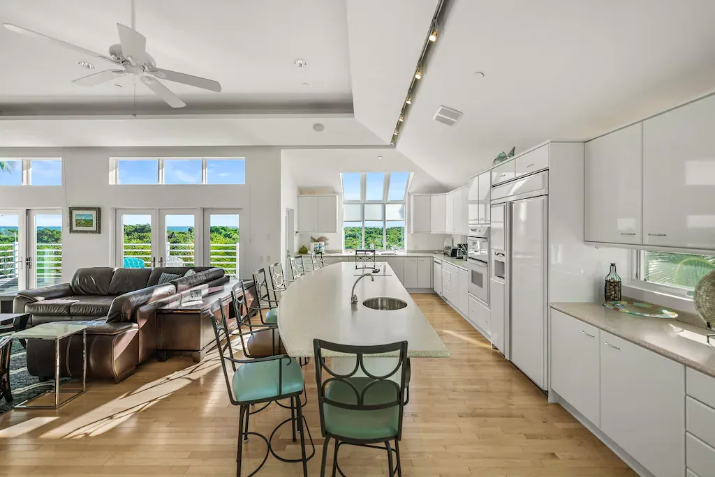Spacious white kitchen with an island, bar stools, and a living room with a brown sofa and outdoor view.