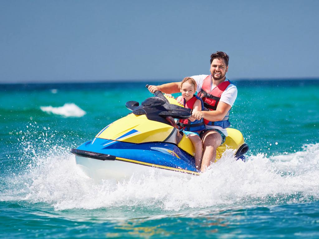 Man and child on a jet ski in turquoise water, both wearing life vests and smiling.