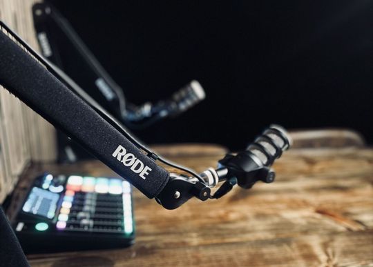 A rode microphone is sitting on a wooden table next to a mixer.