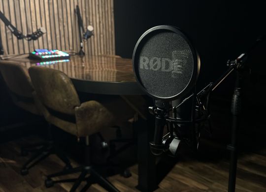 A rode microphone is sitting in a dark room next to a table and chairs.