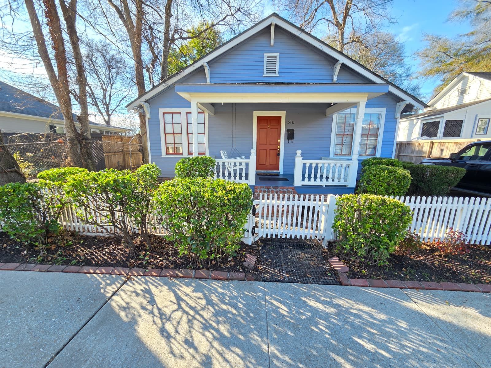 A light blue house with a white front porch, orange door, and a white picket fence in front of manicured bushes.